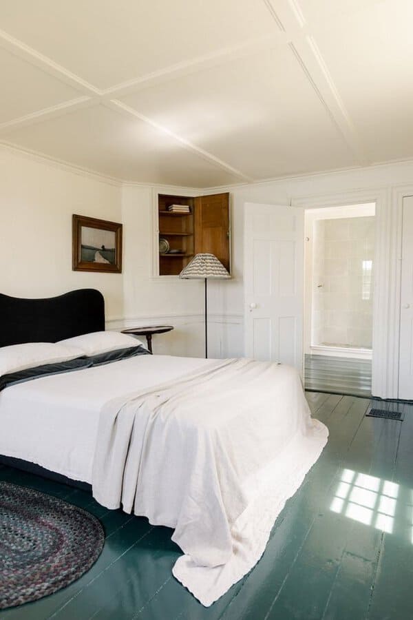 Bedroom with a dark green painted wood floor, white walls, and simple neutral bedding, plus a small braided rug.