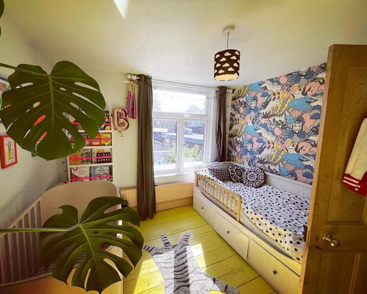 Kids’ room with a bright yellow floor, colorful wallpaper, simple furniture, and a large monstera plant in the foreground.