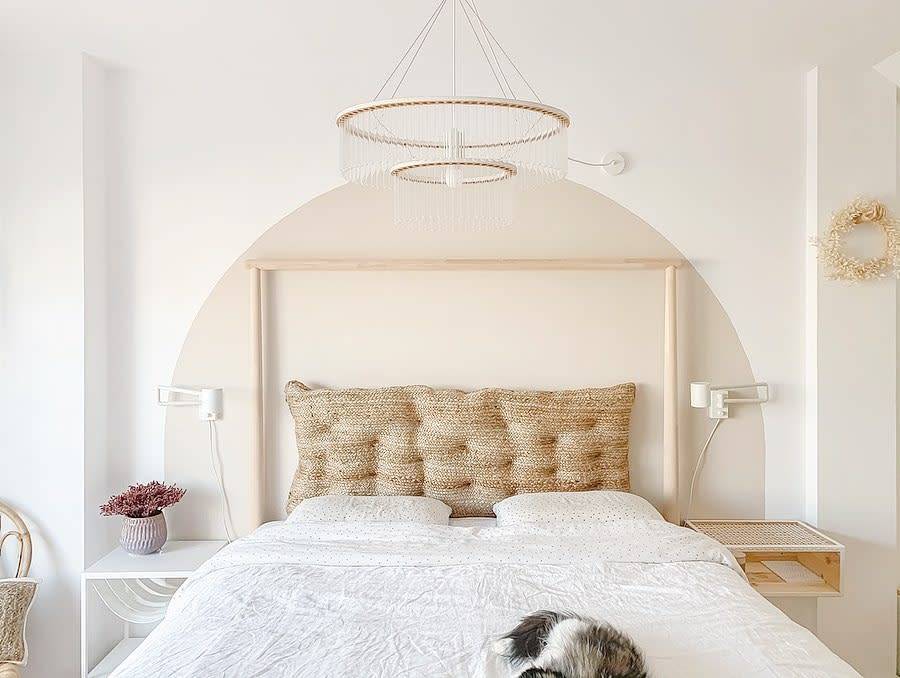 Minimalist bedroom with a woven headboard, white bedding, modern chandelier, and small plant on a side table