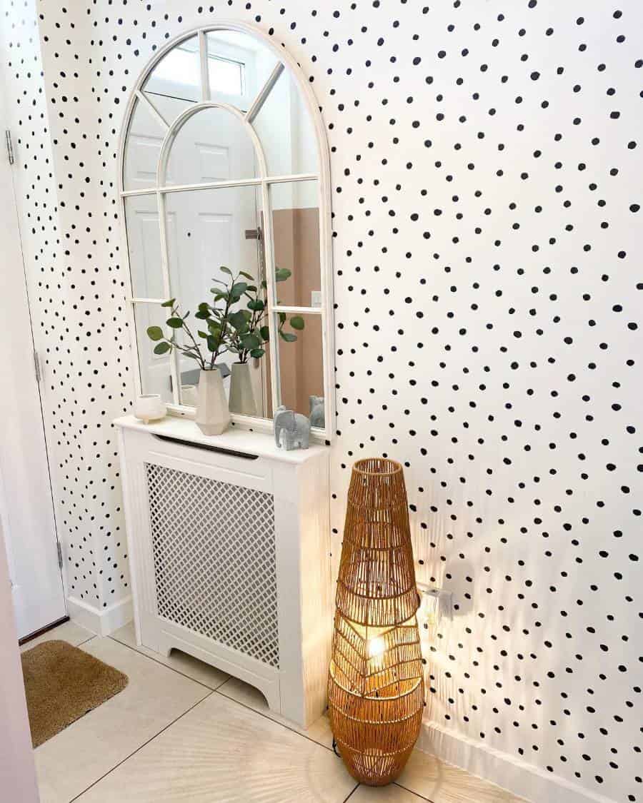 A hallway with a polka dot accent wall, large arched mirror, white cabinet, vase with greenery, and woven floor lamp