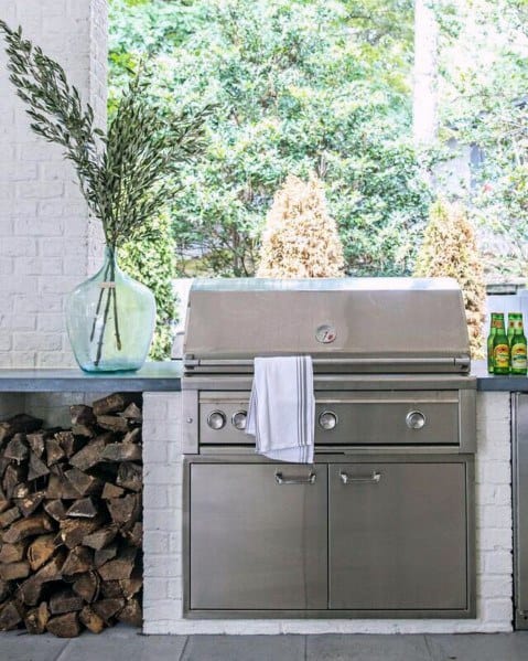 Outdoor patio kitchen with a built-in stainless steel grill, firewood stack, and glass vase with branches