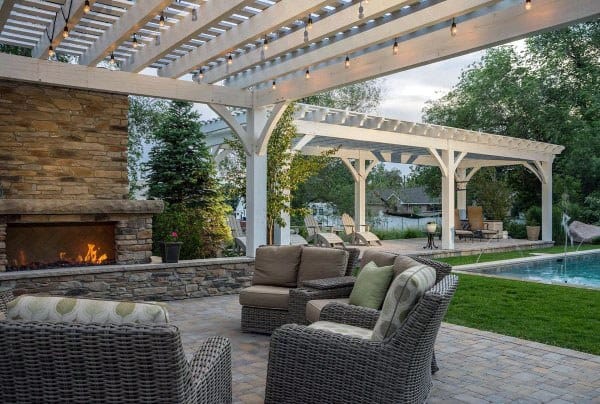 White pergola pavilion with a stone fireplace, wicker seating, and string lights, overlooking a poolside area.