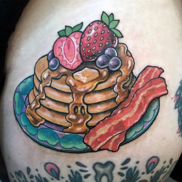 Colorful pancake tattoo featuring syrup, butter, strawberries, blueberries, and crispy bacon on a patterned plate, designed in a bold traditional style.
