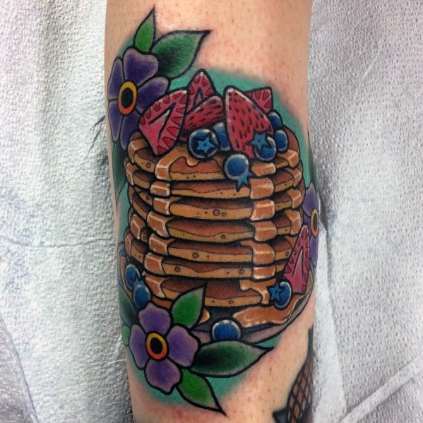 Vibrant pancake tattoo featuring a tall stack with syrup, strawberries, blueberries, and purple flowers, designed in a bold traditional style.