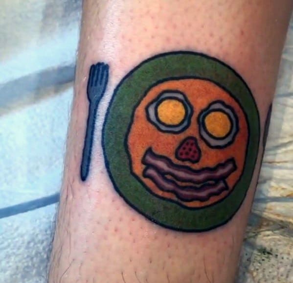 Playful pancake tattoo featuring a breakfast face with eggs as eyes, bacon as a mouth, and a strawberry nose on a green plate with a fork.