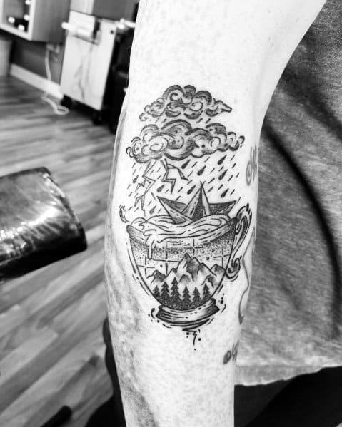 Black ink tattoo of a paper boat in a teacup, with a storm above, lightning, rain, and a landscape inside, symbolizing resilience.
