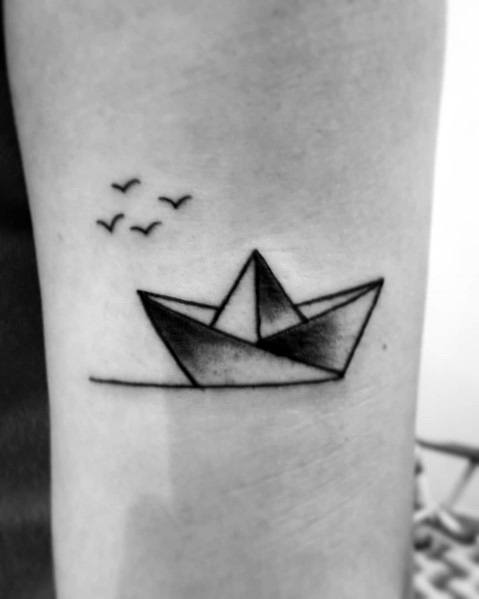 Simple black ink tattoo of a paper boat floating on water with birds flying above, symbolizing adventure, freedom, and serenity.