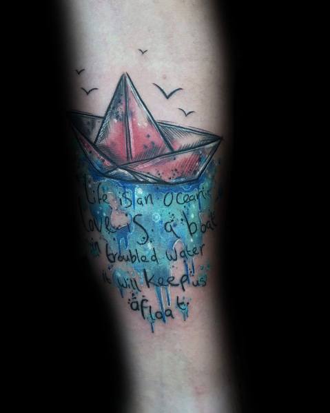 Colorful tattoo of a paper boat on water with a quote beneath, blending blue waves and text, symbolizing resilience and staying afloat in life.
