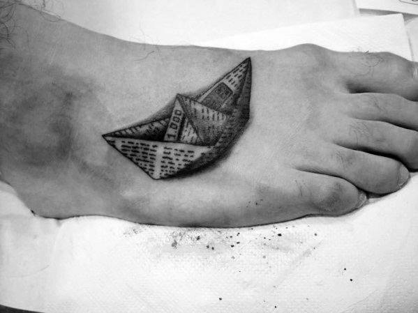 Realistic black ink tattoo of a paper boat made from newspaper on the foot, symbolizing journey, change, and storytelling.