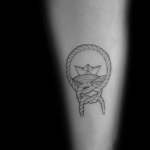 Minimalist black ink tattoo of a paper boat on waves framed by a rope circle, symbolizing adventure, resilience, and nautical themes.