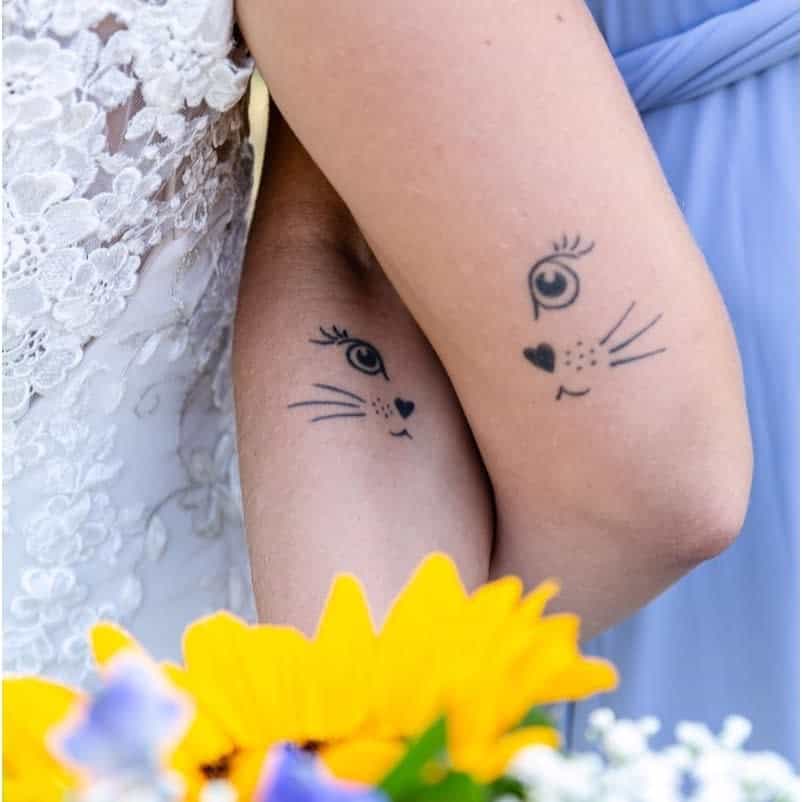 Matching sister tattoos of minimalist cat faces with a heart-shaped nose, whiskers, and expressive eyes