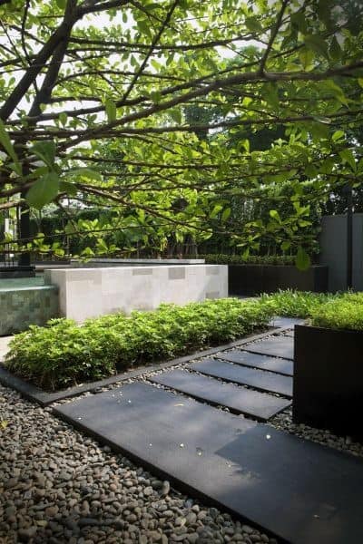 Modern garden with black stone pathway, lush greenery, pebbles, and shaded tree canopy.