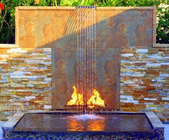 Rustic stone waterfall with a copper backdrop, integrated fire feature, and reflective pool basin.