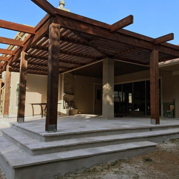 Wooden pergola on a raised stone patio with a fireplace and sliding glass doors.