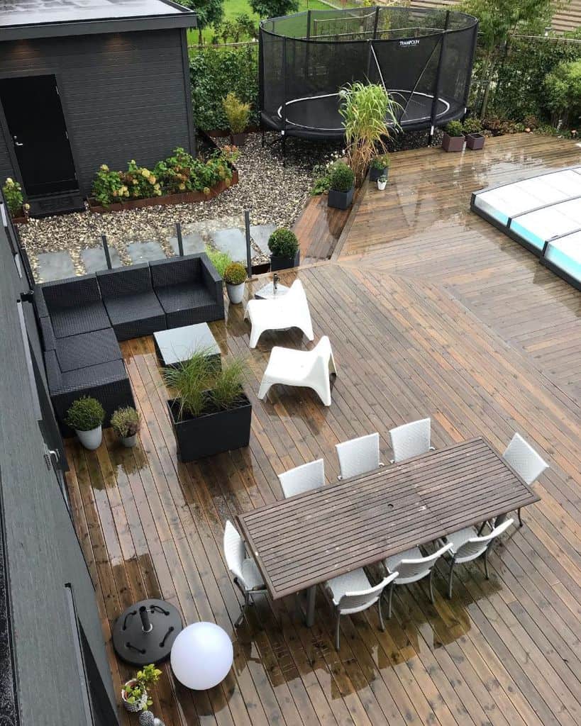 Aerial view of a wet wooden deck with a dining table, chairs, black seating, plants, a trampoline, and a pool cover