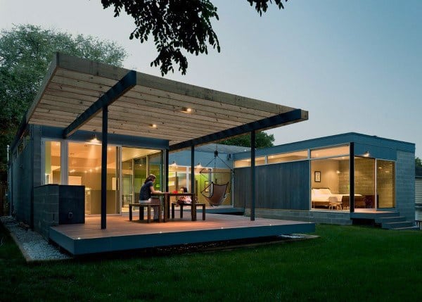 Modern deck with wooden pergola, outdoor dining, and glass-walled home at dusk.