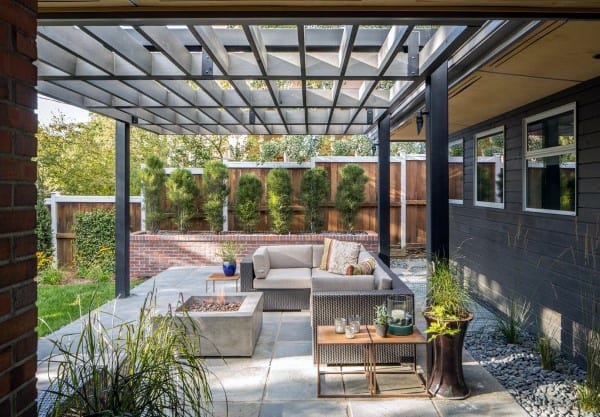 A modern patio features a gray sectional sofa, fire pit, and pergola, all surrounded by lush greenery