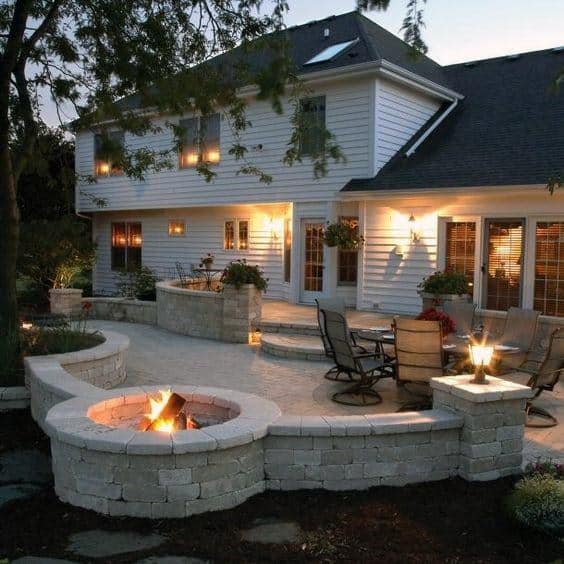 A cozy backyard patio with a fire pit, stone seating walls, comfy chairs, and soft ambient lighting