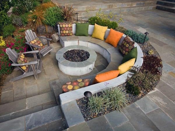 Curved concrete seating with vibrant cushions surrounds a rustic fire pit on a stone patio with wooden chairs
