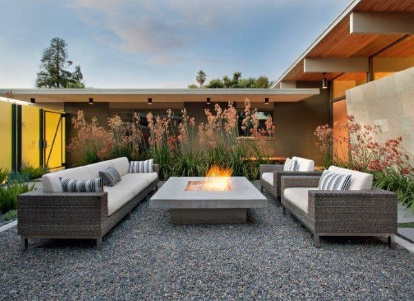 Modern outdoor patio with wicker furniture, fire pit, gravel flooring, and greenery against a sleek house backdrop