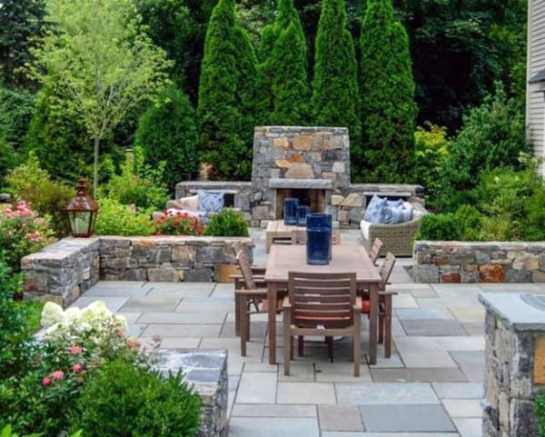 Charming garden patio with a stone fireplace, lush greenery, and a wooden table and chairs in the center