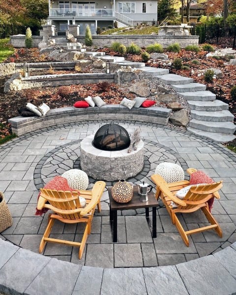 A circular patio with a cozy fire pit, surrounded by chairs, pillows, and steps, with a house and garden gracing the background