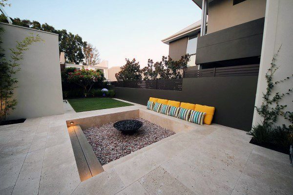 Modern patio with sunken fire pit, pebble ground, and colorful cushions surrounded by lush greenery
