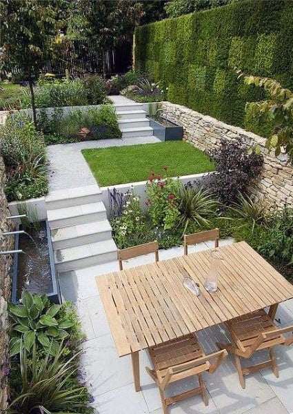 Modern patio garden with a wooden table, surrounded by lush greenery, stone walls, and a tranquil water feature
