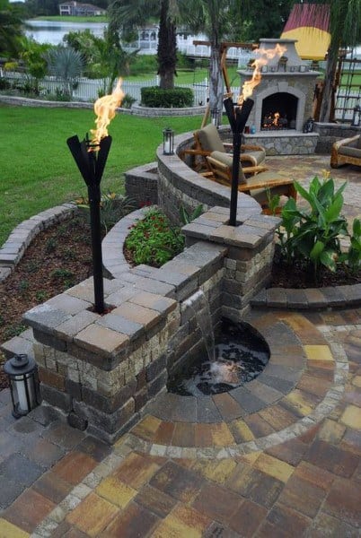 Patio with stone fireplace, torches, water feature, and curved seating in a lush landscaped garden
