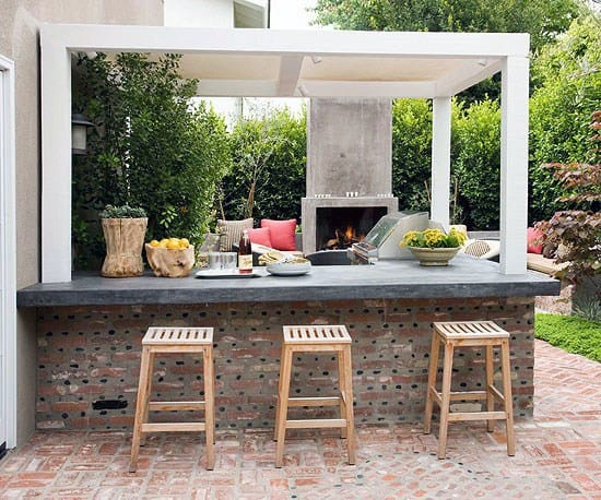 Brick backyard bar with wooden stools and a cozy fireplace surrounded by greenery.