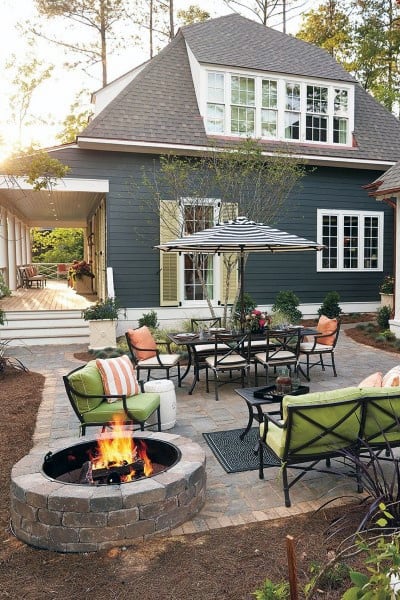 A cozy backyard patio with a fire pit, chair, and striped umbrella beside a dark blue house
