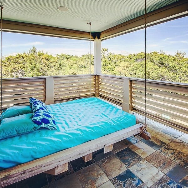 Hanging bed with bright blue bedding on a covered patio overlooking lush greenery.
