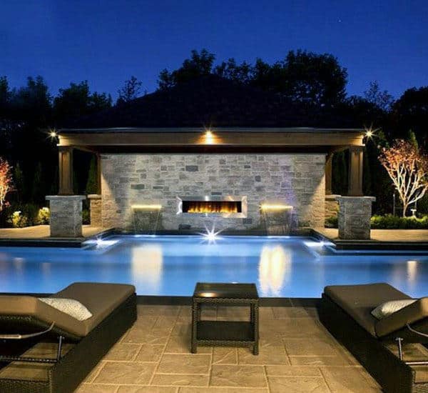 Luxurious outdoor pool area at night with a lit fireplace and lounge chairs