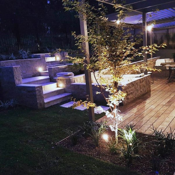 Stone retaining walls with integrated lighting, wooden deck, pergola, and landscaped garden.