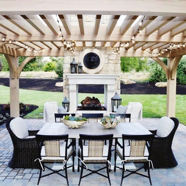 Charming patio with wicker chairs, a dining table, string lights, and a stone fireplace under a wooden pergola
