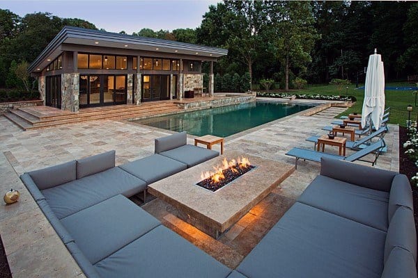 Modern patio with fire pit, gray seating, pool, stone deck, and glass house nestled among trees