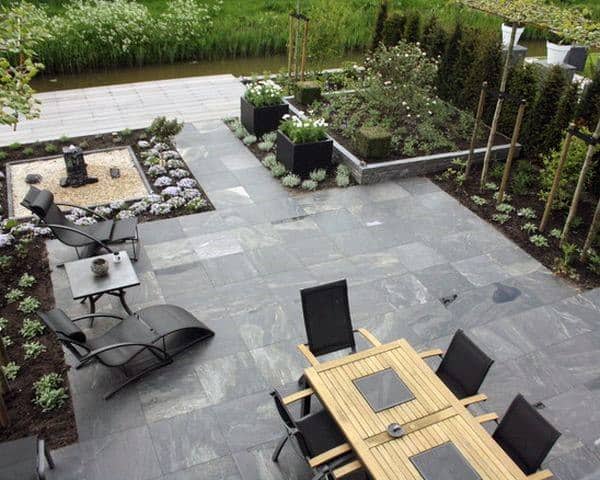 Modern patio with wooden dining set, black loungers, neat landscaping, and a small water feature view