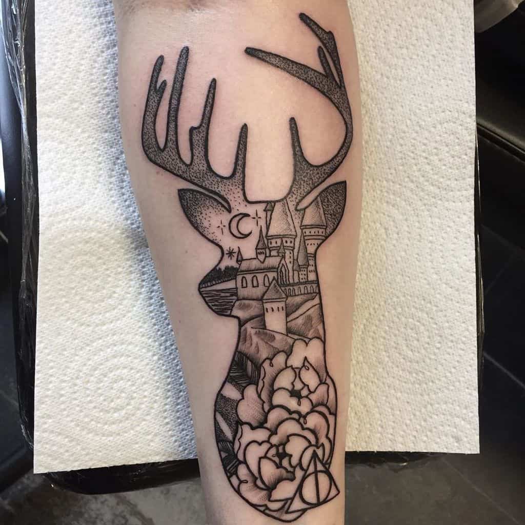 Black ink tattoo featuring a stag silhouette filled with Hogwarts castle, a crescent moon, flowers, and a small Deathly Hallows symbol.
