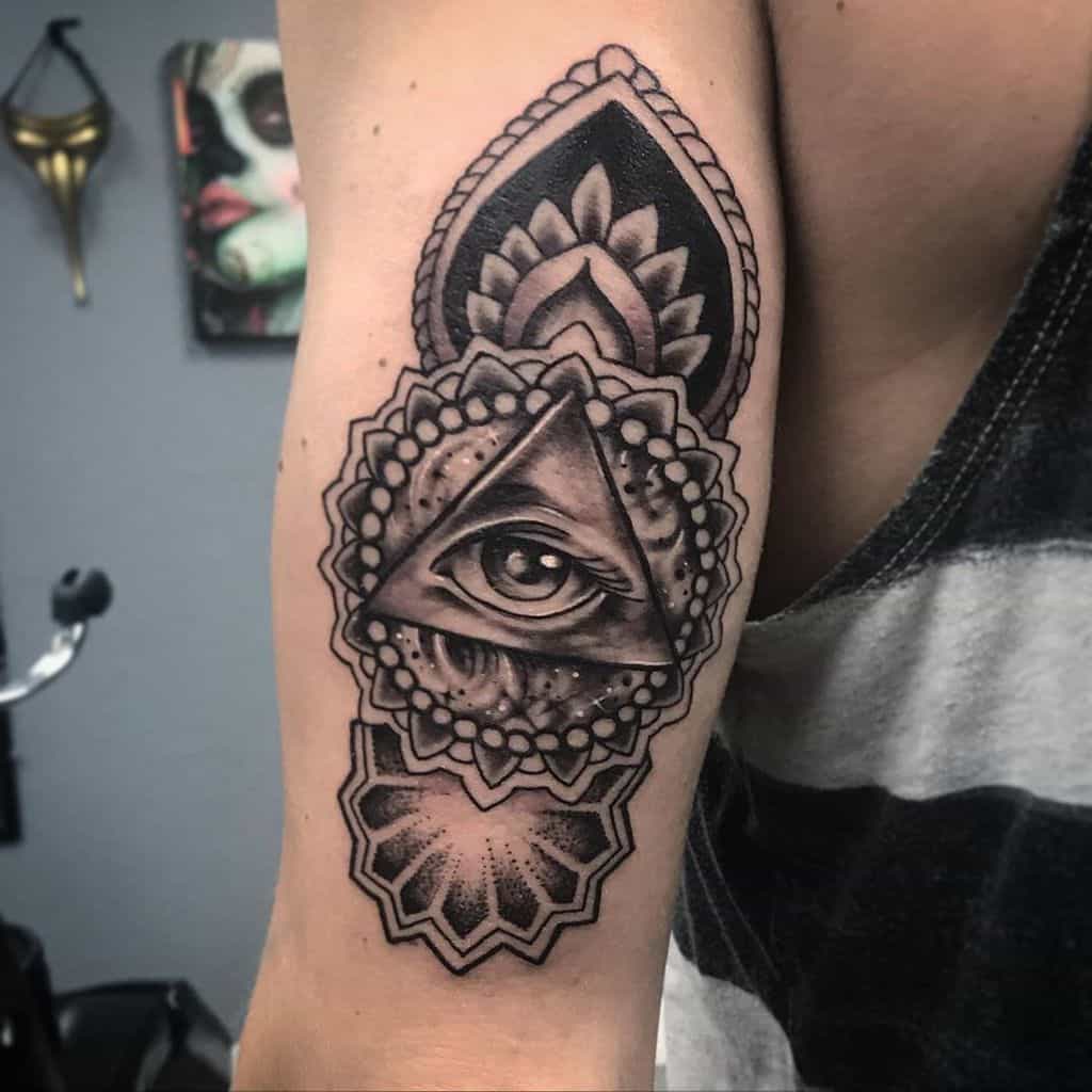 Tattoo of an all-seeing eye inside a triangle, surrounded by intricate mandala and ornamental patterns, inked in black and grey on the upper arm.