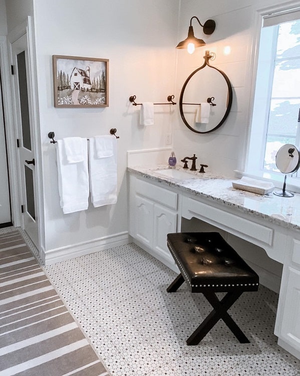 A modern farmhouse bathroom with a round mirror, striped rug, and a cushioned stool by the vanity