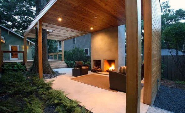 Modern pavilion with a wooden ceiling, minimalist fireplace, and cozy seating area, surrounded by greenery.