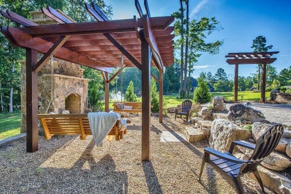Charming pergola pavilion with wooden swings, stone fireplace, and Adirondack seating in a natural landscape.