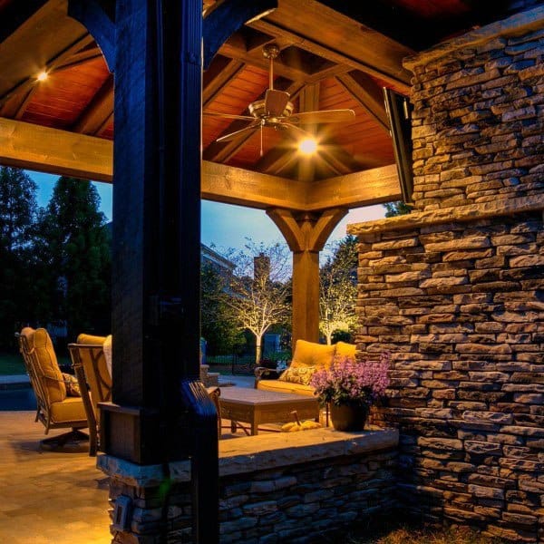 Rustic stone pavilion with wooden beams, ceiling fan, and comfortable seating illuminated by soft lighting.