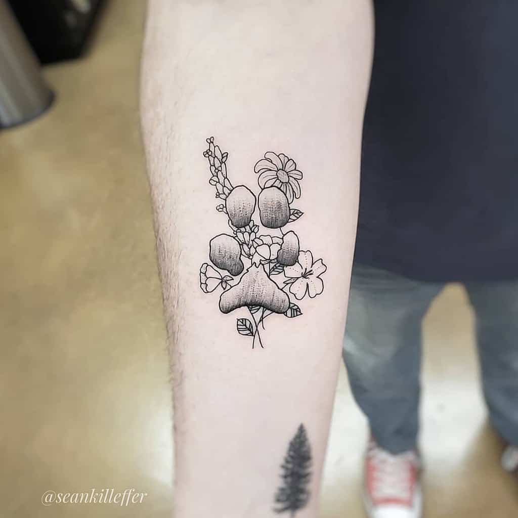A tattoo on the forearm featuring a delicate design of flowers including lilies and other floral elements, with fine line detailing and shading. The tattoo has a soft and minimalistic feel, focusing on the natural beauty of the flowers.