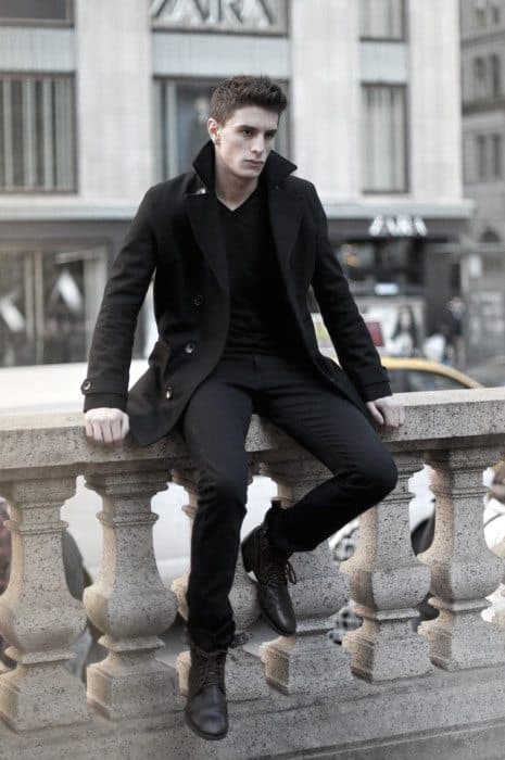 A man in a men’s peacoat sits on a stone railing in the city, gazing to the side with a serious expression and winter style