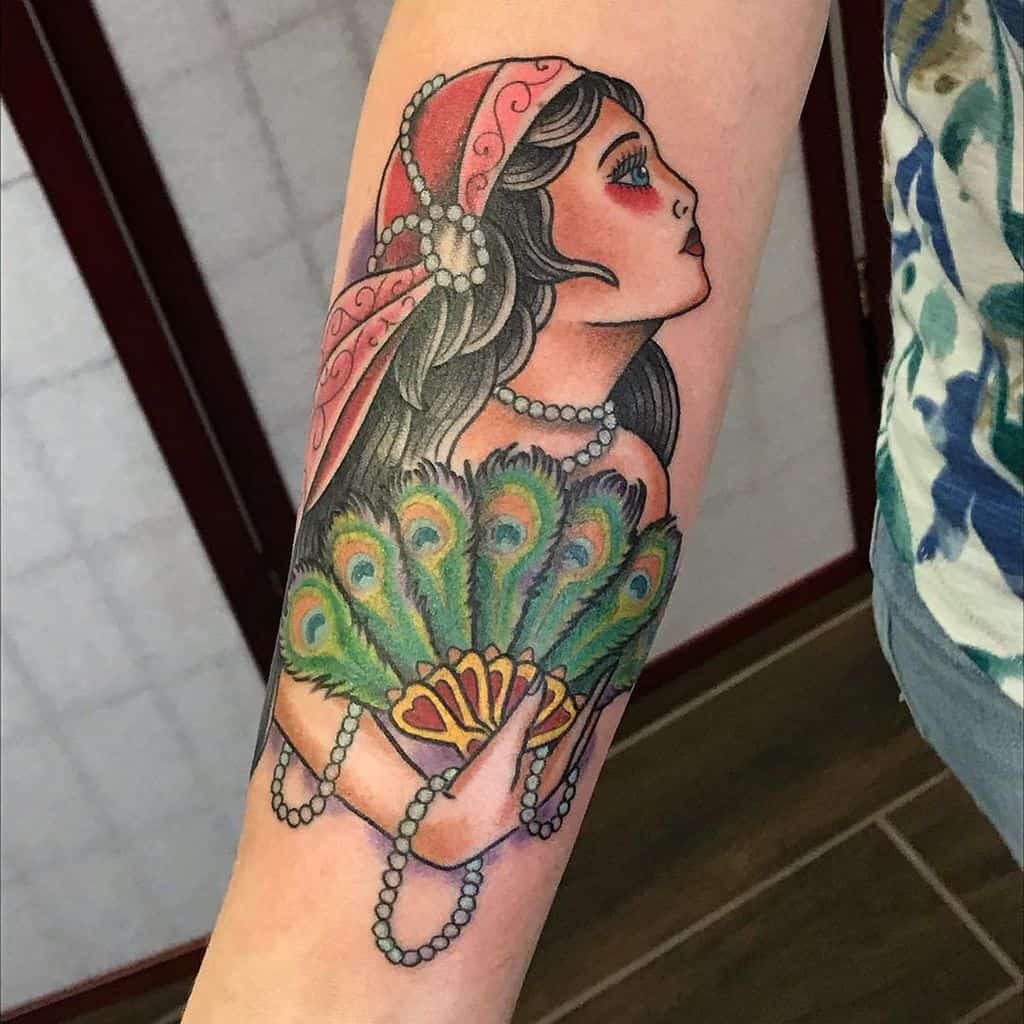 Traditional tattoo of a gypsy woman with a red headscarf, pearl jewelry, and a vibrant peacock feather fan on the forearm.