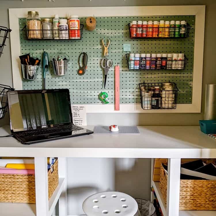 Craft a workspace with a white desk, laptop, pegboard featuring tools, paints, and storage baskets