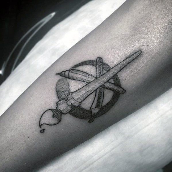 A forearm tattoo featuring a crossed pencil, paintbrush, and pen elegantly enclosed within a circle