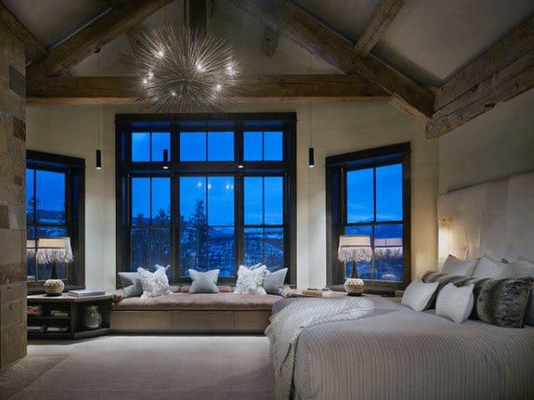 Rustic bedroom with large windows, wooden beams, cozy seating, and starburst chandelier.