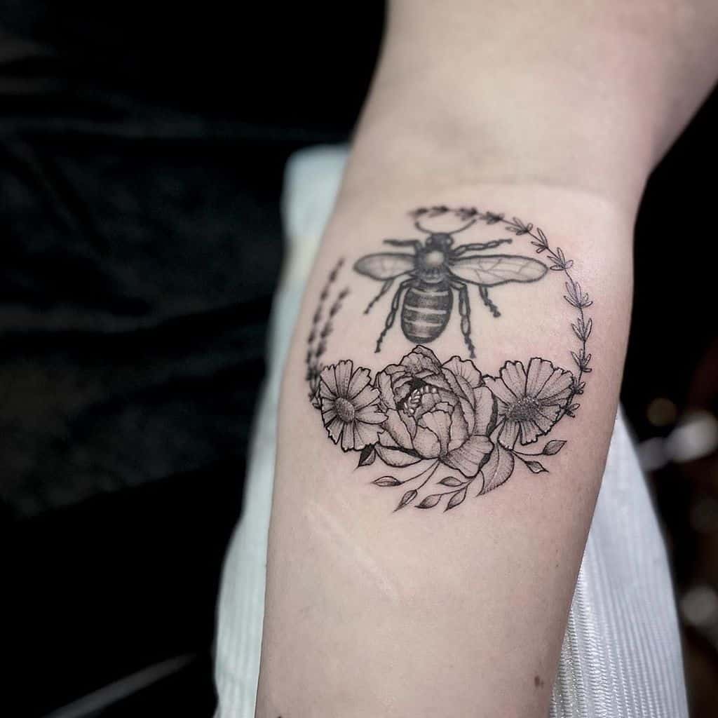 A black and white tattoo of a bumblebee surrounded by a circular wreath of flowers and leaves, placed on the forearm.
